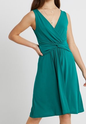 Vrouw die een mouwloze teal jurk draagt met een V-hals, een gedraaide knoop in de taille en gelaagde gouden kettingen, poserend met één hand op haar heup.