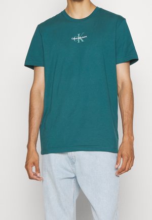 Homme portant un t-shirt Calvin Klein Jeans manches courtes couleur sarcelle et un jean bleu clair, debout devant un fond uni.