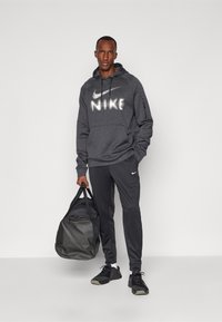 Šedá mikina Nike s bielym logom, zodpovedajúce šedé nohavice a čierne tenisky. Drží čiernu športovú tašku. Biely pozadí.
