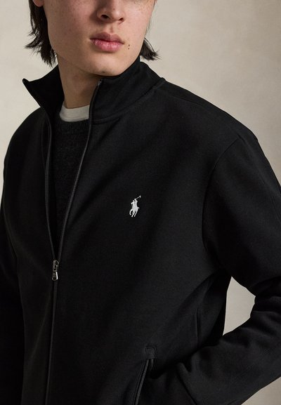 Jeune personne portant une veste noire partiellement zippée avec un logo blanc de joueur de polo sur la poitrine, debout devant un fond uni.