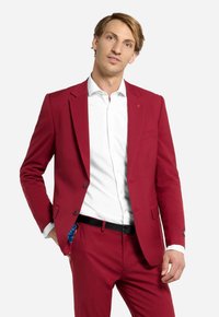 Completo rosso con vestibilità slim, caratterizzato da una giacca a un solo petto, rever a punta e una camicia bianca sotto. Accento blu visibile nella tasca.