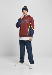 Burgunderfarbener Hoodie mit gelben und blauen Akzenten, "STARTER"-Logo, kombiniert mit blauen Jeans und weißen Sneakers. Modell trägt eine passende Mütze.