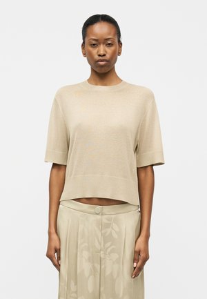 Γυναίκα που φοράει ένα μπεζ πουλόβερ με κοντά μανίκια και παντελόνι ψηλόμεσο, ανοιχτό μπεζ με ένα διακριτικό floral μοτίβο, στέκεται μπροστά σε ένα απλό φόντο.