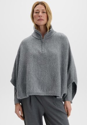 Frau mit blonden Haaren, die ein hellgraues gestricktes Poncho mit Reißverschluss über langärmligen Shirts und dunkelgrauen Hosen vor einem einfarbigen Hintergrund trägt.