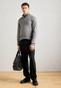 Gray zip-up sweater with long sleeves, black trousers, and black shoes. Holding a black leather bag, standing on wooden floor against a light backdrop.