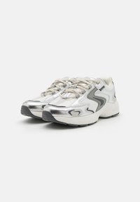 Scarpe da ginnastica argento realizzate con un mix di materiali testurizzati, con lacci prominenti, suole supportive e linee di accento curve in grigio.