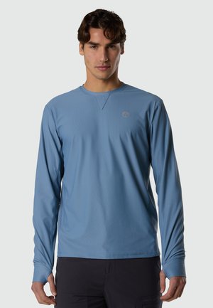 Mann trägt ein hellblaues Langarmhemd mit einem kleinen Logo auf der Brust, steht vor einem einfarbigen Hintergrund.