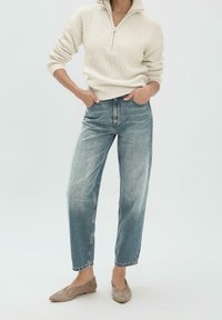 Frau trägt einen cremefarbenen Strickpullover mit einem halben Reißverschluss, ausgebleichte blaue High-Waist-Jeans und beige Wildleder-Ballerinas, mit den Händen in den Taschen.