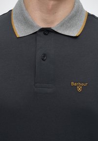 Polo gris foncé avec col à motif orné de bordure moutarde, deux boutons noirs et logo Barbour jaune sur la poitrine.