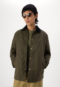 Giacca verde oliva con colletto in velluto a coste nero, bottoni a pressione e tasche laterali, indossata sopra un maglione verde a trama.