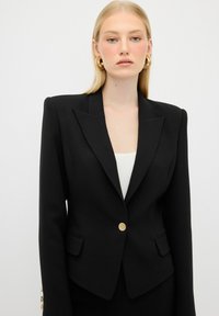 Blazer nero su misura con rever affilato, chiusura con un singolo bottone dorato e due tasche frontali. Tessuto liscio e forma strutturata.