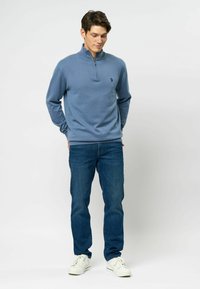 Világoskék poliészter pulóver fél cipzárral a gallérnál, amelyen egy kis logó található, kék farmernadrággal és fehér sportcipővel párosítva.