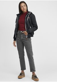 Black hooded jacket with white zipper, maroon ribbed sweater, gray high-waisted jeans, and tan platform sneakers with black accents.
