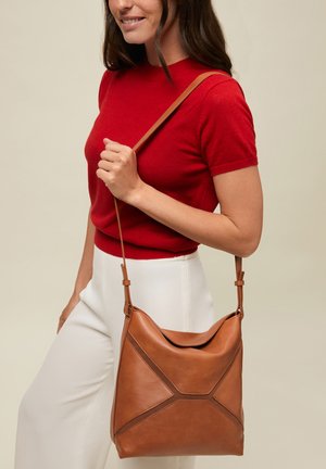 Kobieta w czerwonym topie z krótkim rękawem i białych spodniach, trzymająca brązową skórzaną torebkę na ramię z długim paskiem.