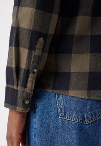 Cămașă din flanel în carouri maro și albastru închis, cu mansete cu nasturi, alături de blugi de denim albastru estompat, având un buzunar pe spate. Material din amestec de bumbac.