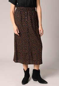 Femme portant une jupe midi noire avec de petits pois rouges, à boutons devant, taille élastique et bottines noires.