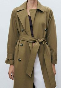 Manteau trench kaki avec un design à double boutonnage, des poches avant et une ceinture à nouer. Fabriqué en tissu lisse, avec un col en V et des manches longues.