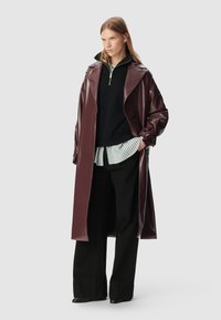 Woman standing wearing a long glossy dark brown coat, black wide-leg pants, black zip-up sweater, and striped shirt layered underneath.