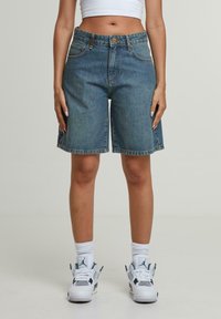 Zils džinsu šortu modelis ar izbalējušu toņu, piedāvā klasisku piecu kabatu dizainu un taisnu kāju griezumu, valkāti ar baltām sporta apaviem.