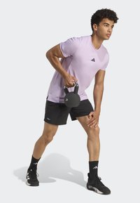 Kettlebell negra sostenida por una persona con una camiseta deportiva de color púrpura claro y pantalones cortos negros, con calcetines y zapatos negros, en una posición lista para hacer ejercicio.