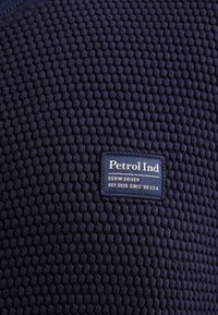 Marin textil med ett våffelmönster. Har en blå etikett med vit text: "Petrol Ind Denim Driven Ref.5026 Sedan '89 U.S.A."