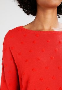 Roter Strickpullover mit weitem Ausschnitt und strukturierten, runden Noppen-Details. Weicher Stoff und lockere Passform.