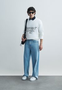 White sweatshirt with "Emporio Armani" logo, dark collared shirt underneath, light blue wide-leg jeans, and white sneakers, completed with sunglasses.