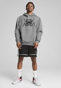 Sweat à capuche gris avec le logo "PUMA" noir, shorts noirs avec des accents blancs et baskets multicolores ; le modèle se tient devant un fond neutre.