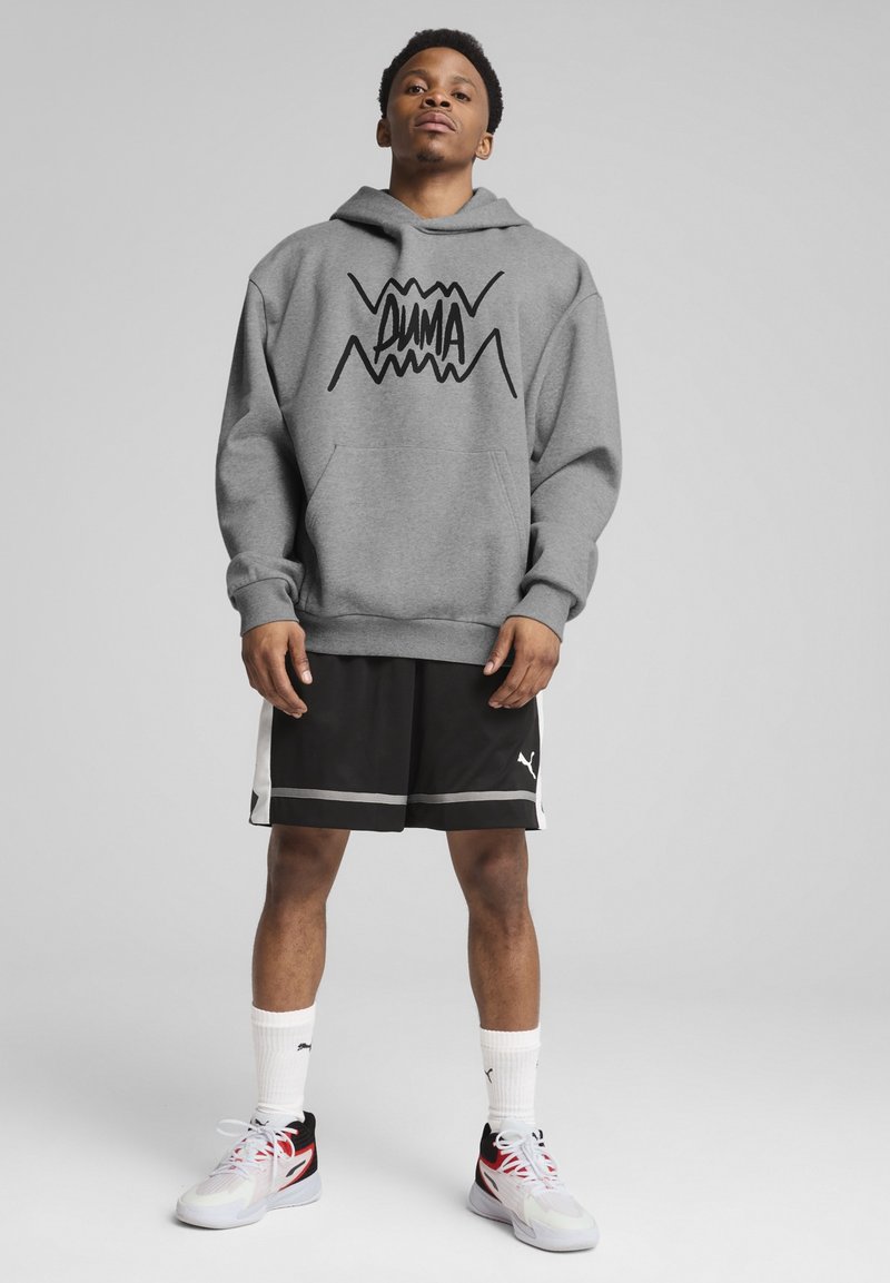 Sweat à capuche gris avec le logo "PUMA" noir, shorts noirs avec des accents blancs et baskets multicolores ; le modèle se tient devant un fond neutre.