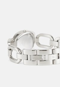 Montre-bracelet en acier inoxydable avec une finition polie, dotée d'un cadran rectangulaire et d'un bracelet à maillons épais. Nom de la marque "GUESS" affiché sur les maillons métalliques.