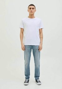 Ung mann står med ansiktet vendt fremover iført en enkel hvit t-skjorte, lyseblå jeans og svarte joggesko mot en ensfarget bakgrunn.