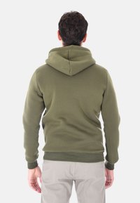 Uomo che indossa un hoodie verde oliva e pantaloni grigi chiari, in piedi di spalle contro uno sfondo bianco uniforme.