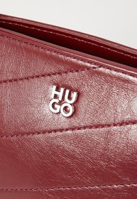 Borsa in pelle bordeaux con texture trapuntata, accentuata dal logo "HUGO" in argento e dettagli impunturati lungo i bordi.