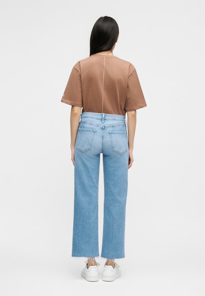 Γυναίκα με μακριά σκούρα μαλλιά, φορώντας καφέ κοντομάνικη μπλούζα, ανοιχτόχρωμα μπλε ψηλόμεσα κατεβασμένα τζιν και λευκά αθλητικά παπούτσια, στέκεται με την πλάτη προς τα εμπρός.