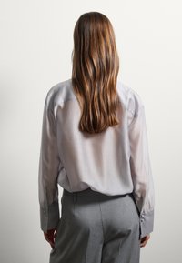 Camicia grigia chiara a maniche lunghe con una texture morbida e liscia. Presenta polsini sartoriali con dettaglio a bottone, indossata con pantaloni grigi.