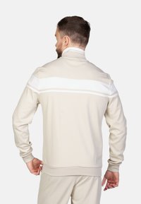 Giacca beige con zip e colletto alto. Presenta strisce bianche sulle spalle, maniche lunghe e polsini a coste. Tessuto con una superficie liscia.