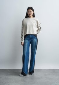 Žena stojí opretá o jednoliatu stenu, oblečená v krémovom top so vzorovanými kvetinovými rukávmi a vo vysokým pásom modrých a čiernych zvonových džínsach so striebornými špicatými topánkami.