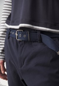 Marina bomullshängselbyxor med ett strukturerat blått bälte och spänne i silvermetall. Toppdelen är marinblå med vita ränder längs ärmarna.