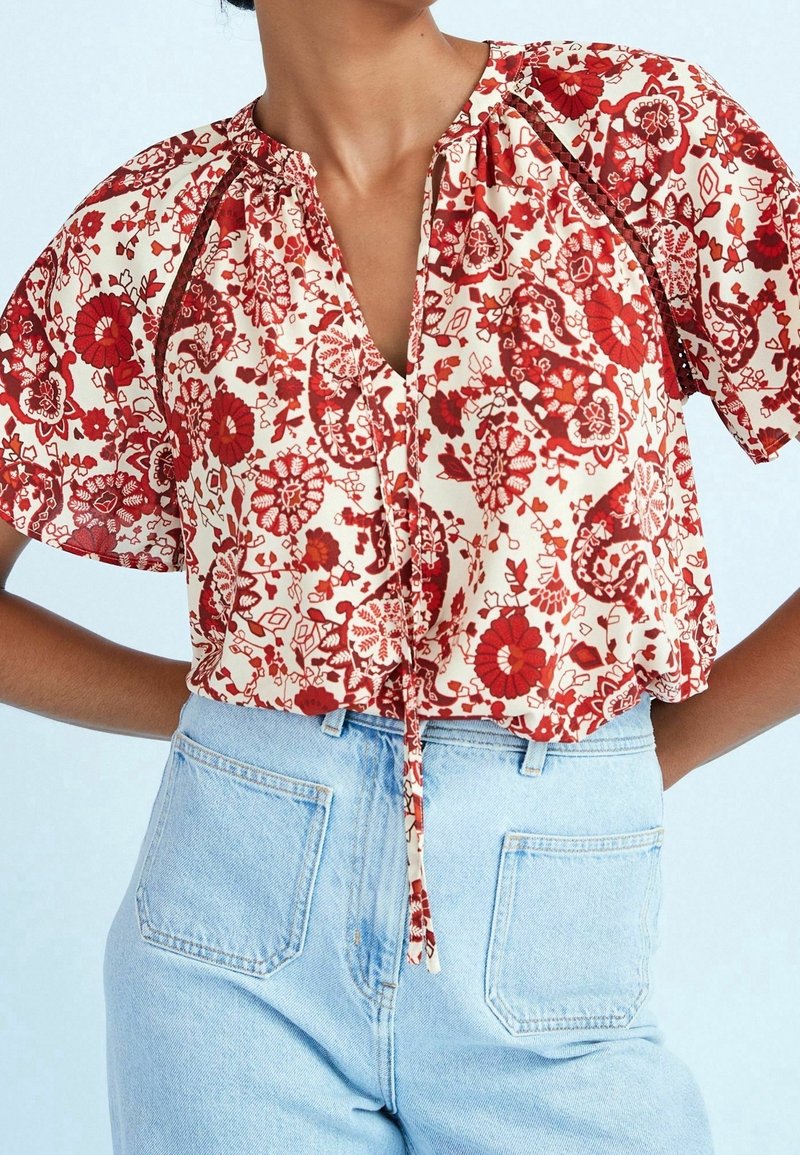 Rode bloempatroon blouse met een V-hals, pofmouwen en strikdetail in de taille, gecombineerd met lichtblauwe high-waisted jeans.