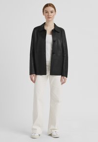Schwarze Lederjacke mit Kragen, zwei vorderen Knöpfen und einer glatten Textur, kombiniert mit einem weißen Oberteil und cremefarbenen, weitgeschnittenen Hosen.