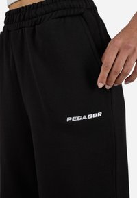 Black sweatpants with an elastic waistband, featuring side pockets and a white embroidered "PEGADOR" logo on the thigh. Smooth texture.