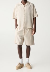 Chemise à manches courtes beige clair avec fermeture à boutons, assortie à un short de la même couleur. Portée avec des chaussures beiges et des chaussettes blanches aux chevilles. Design décontracté.
