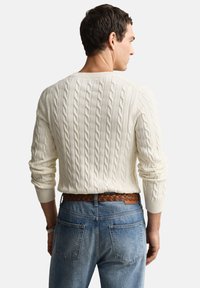 Kreminės spalvos megztinis su pynimais, apvalia apykakle, derinamas su šviesiai mėlynais džinsiniais ir ruduoju austiniu diržu, pabrėžiant tekstūruotus raštus bei laisvą stilių.
