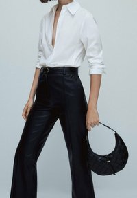 Chemise blanche à boutons et pantalon noir taille haute avec une finition brillante. Sac à main noir matelassé avec une poignée courbée.