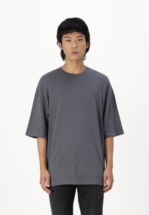 T-shirt oversize gris foncé en coton doux, avec un col rond et des manches courtes, associé à un jean noir. Texture lisse, design minimal.