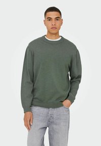 Groene gebreide trui met een ronde hals, lange mouwen en ribbels aan de mouwen. Draag over een wit shirt en gecombineerd met lichtgrijze denim jeans.