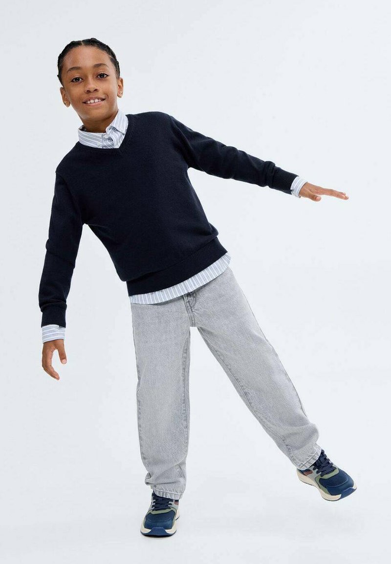 Pulover navy cu decolteu în V peste o cămașă cu guler în dungi albastre deschise, asociat cu blugi gri deschis și adidași albaștri.