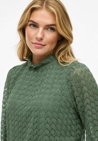 Groene kantblouse met een textuurpatroon, hoge rimpelige halslijn en lange semi-doorzichtige mouwen. Zachte stof met een delicaat ontwerp.