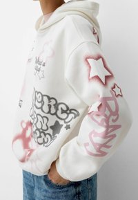 Felpa bianca con cappuccio, caratterizzata da disegni in spray rosa e grigio, stelle e motivi di nuvole. I polsini sono a coste e il tessuto appare morbido.