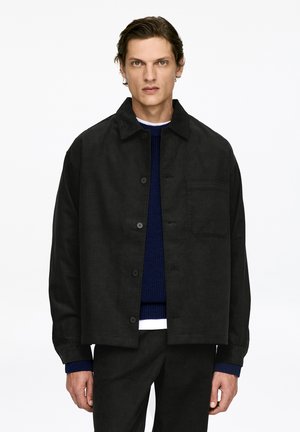 Black corduroy jacket with a button closure, chest pocket, and folded collar, worn over a blue ribbed sweater.